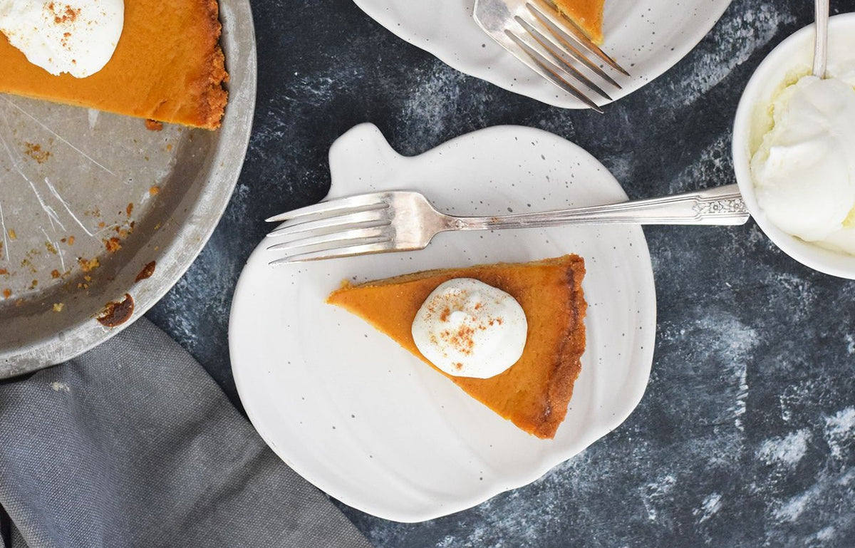 Slice of pumpkin pie with whipped cream on a white plate beside a larger pie