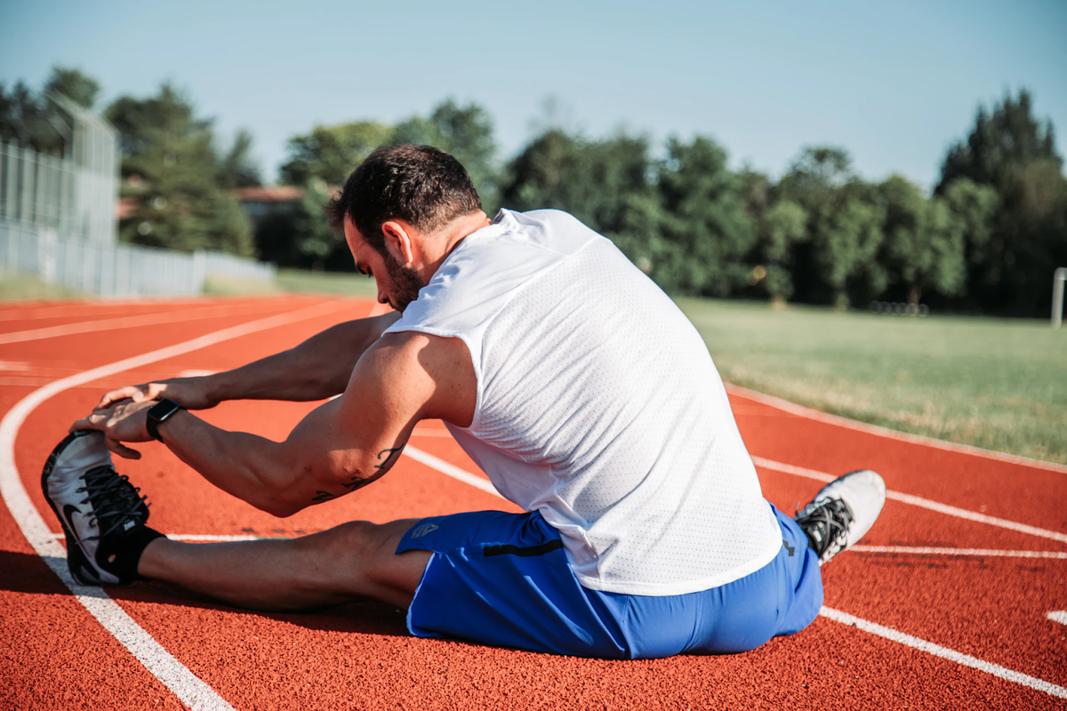 Athlete stretches on a red running track wearing a white sleeveless shirt and blue shorts.
