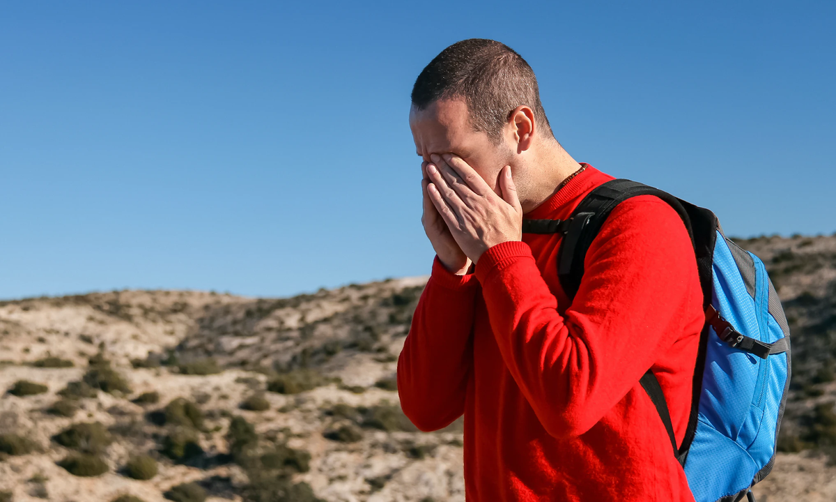 Hiker in a red shirt with a blue backpack shields his eyes outdoors in sunny rocky terrain