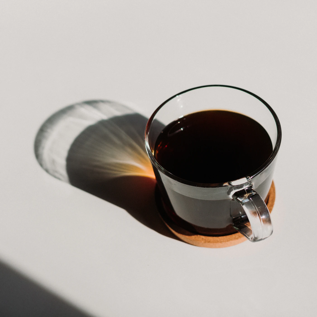 Clear glass cup of black coffee on a cork coaster cast in a soft shadow.