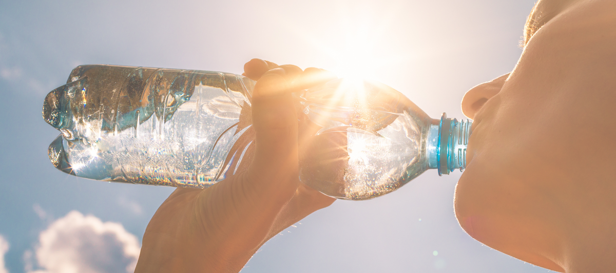 Close up of a hand gripping a clear water bottle as sunlight flashes behind it in a bright outdoor scene.