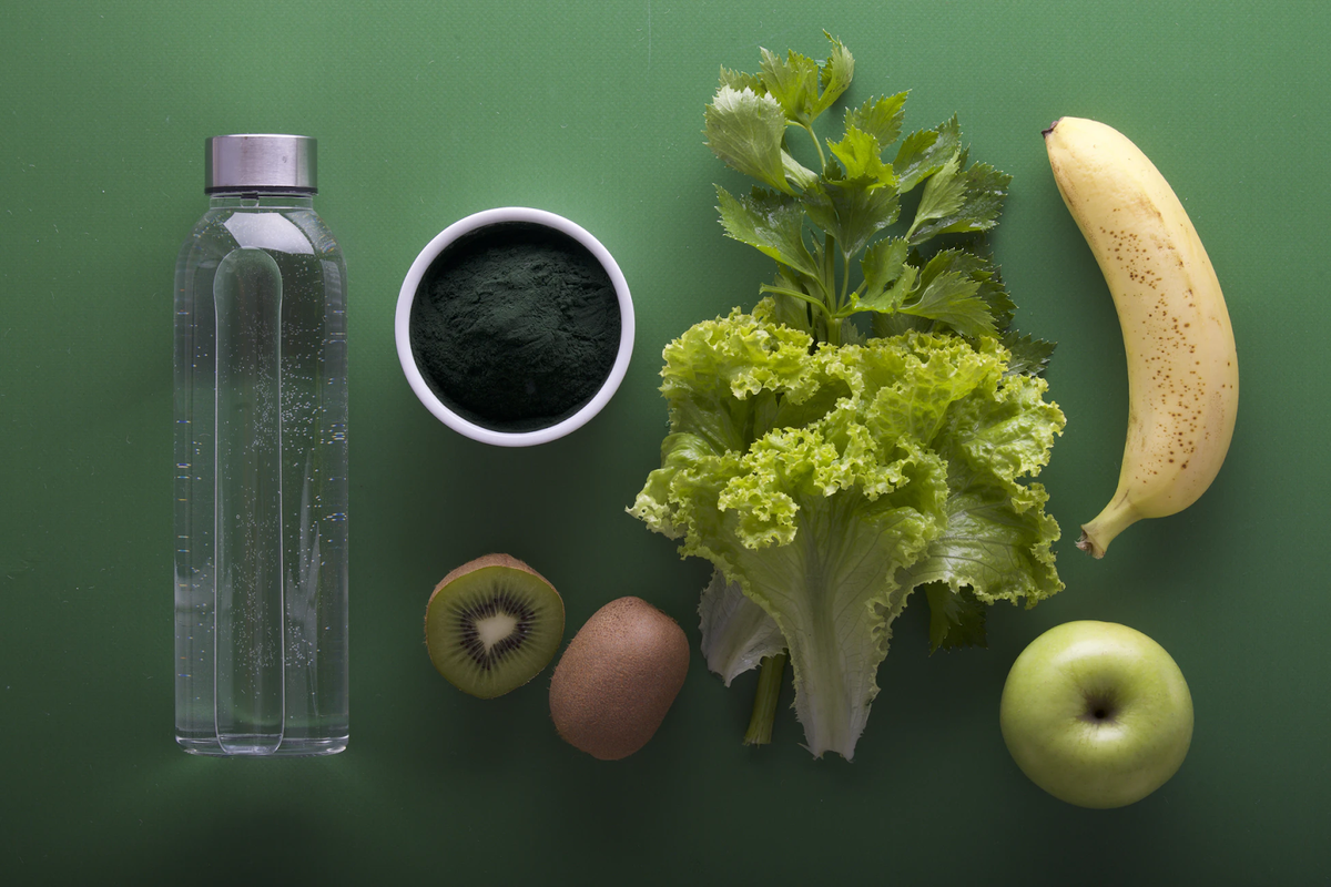 Flat lay of hydration friendly foods with a water bottle, bowl of powder, celery and leafy greens plus banana kiwi and apples on a green background.