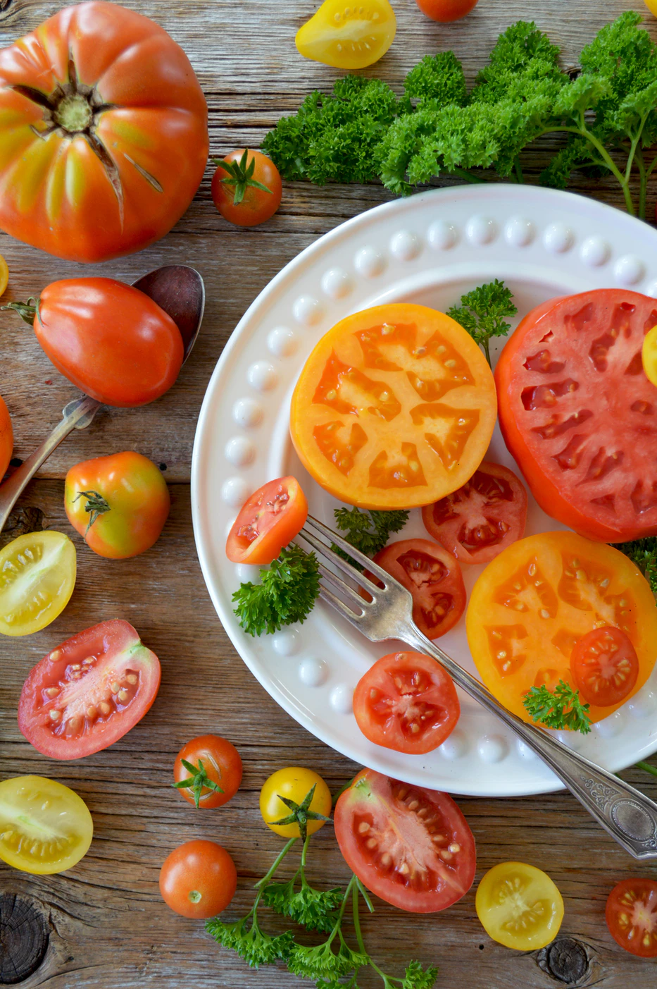 Top view of keto friendly produce with a plate of sliced tomatoes, leafy greens and parsley arranged on a wooden surface with an apple nearby.