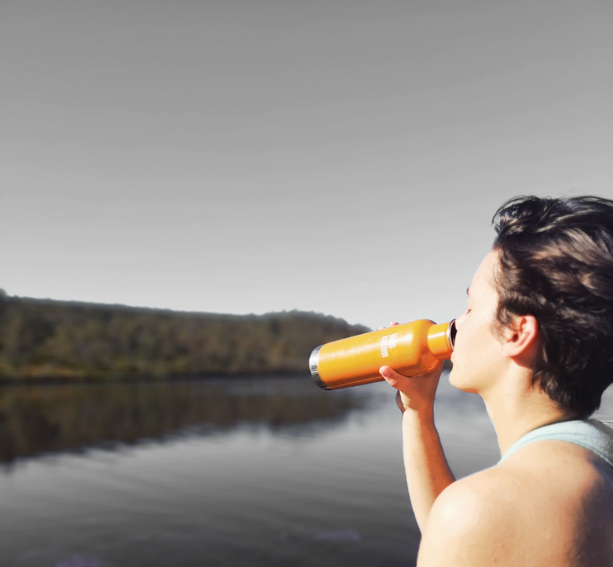 Person drinking from an orange sports bottle by a lake on a sunny day.