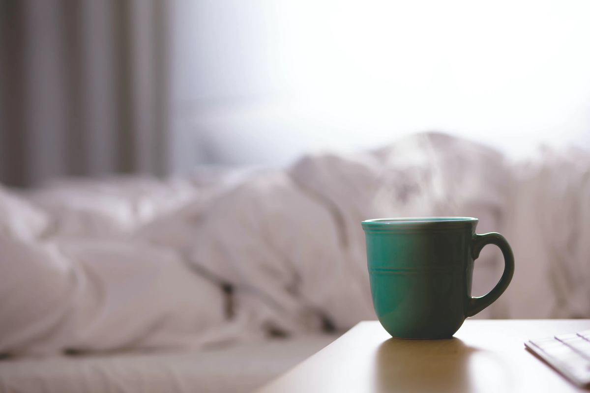 Teal green mug sits on a wooden bedside table with a soft blurred bed in the background.