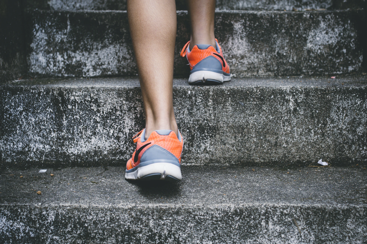 Athlete's feet in orange and gray running shoes step up a set of rough concrete stairs.
