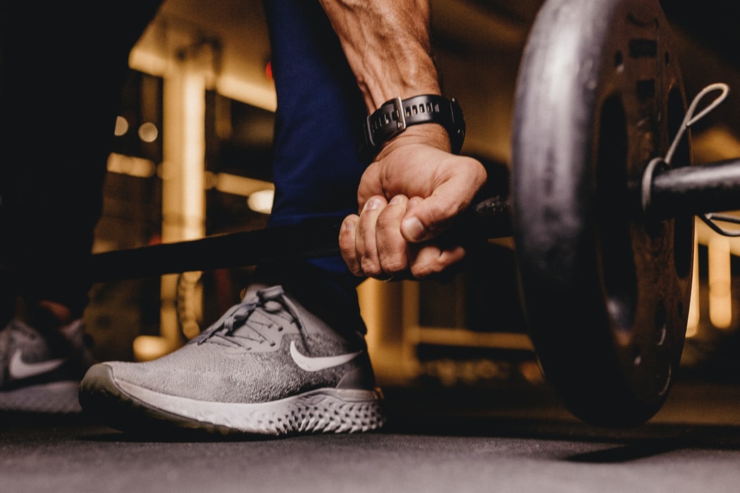 Close up of a lifter's hand gripping a barbell on a gym floor with a gray running shoe in the background