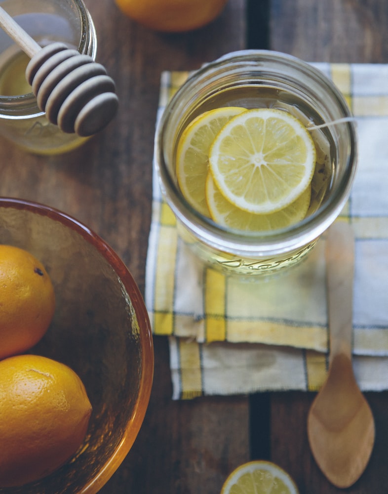Lemon water in a glass jar with lemon slices, honey dipper, and a bowl of oranges on a wooden table