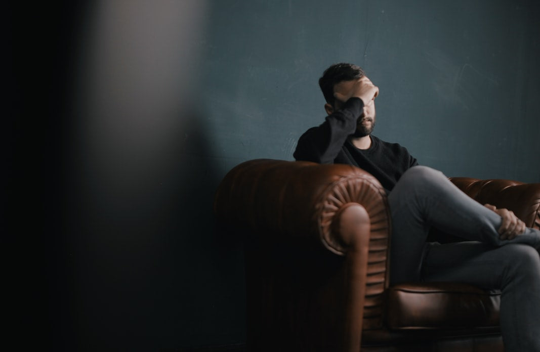 Man sitting on a brown leather sofa, holding his head in a somber room