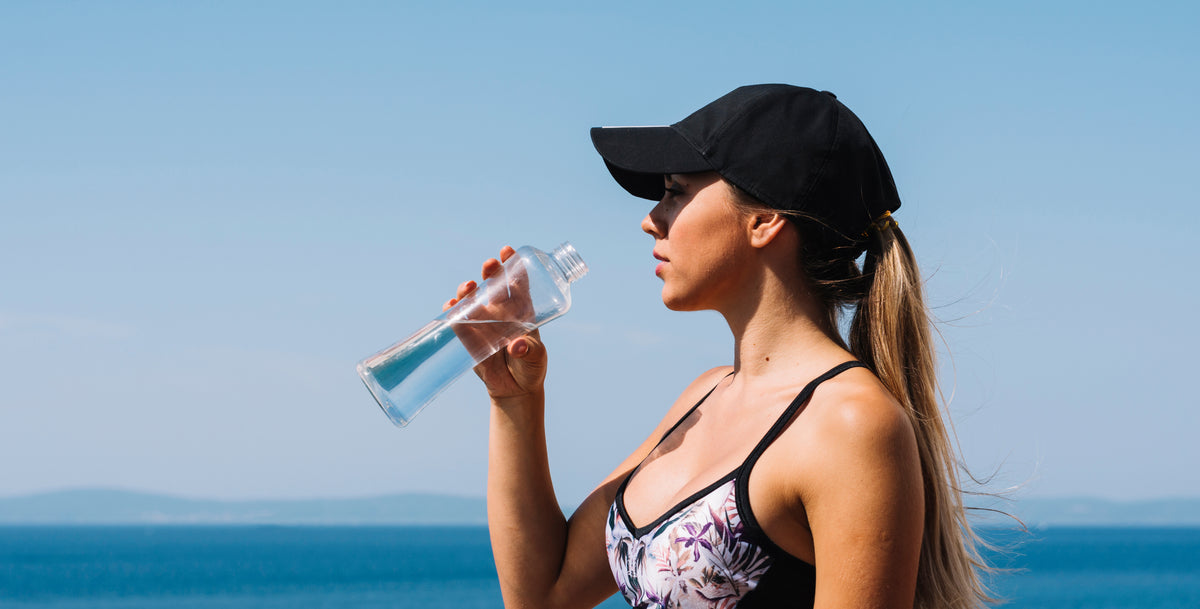 Woman in a black cap drinks water from a bottle with the ocean in the background.