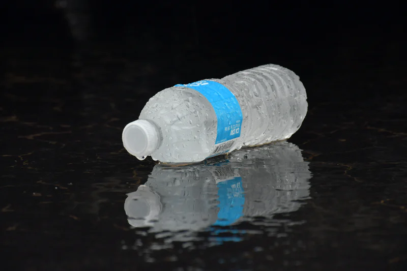 Crushed clear water bottle with blue label lying on a wet dark surface with its reflection