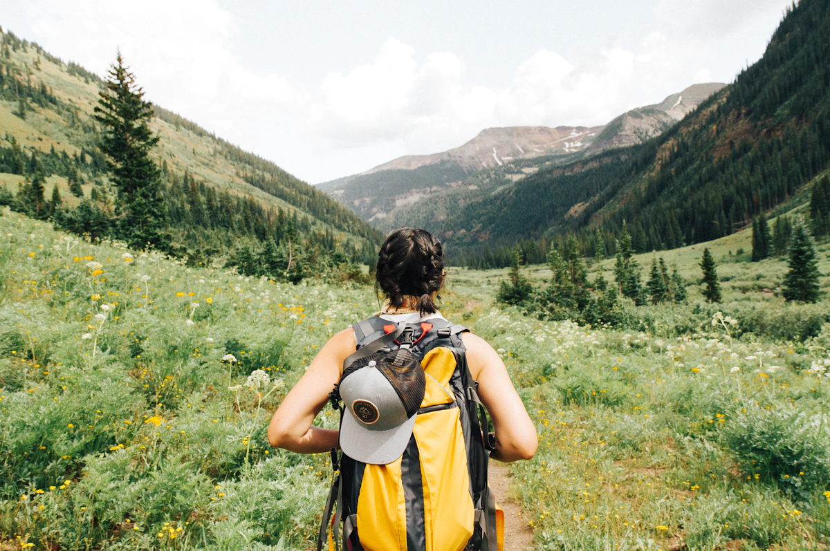 Back view of a hiker wearing a yellow and black backpack hiking through a meadow of wildflowers toward mountains.