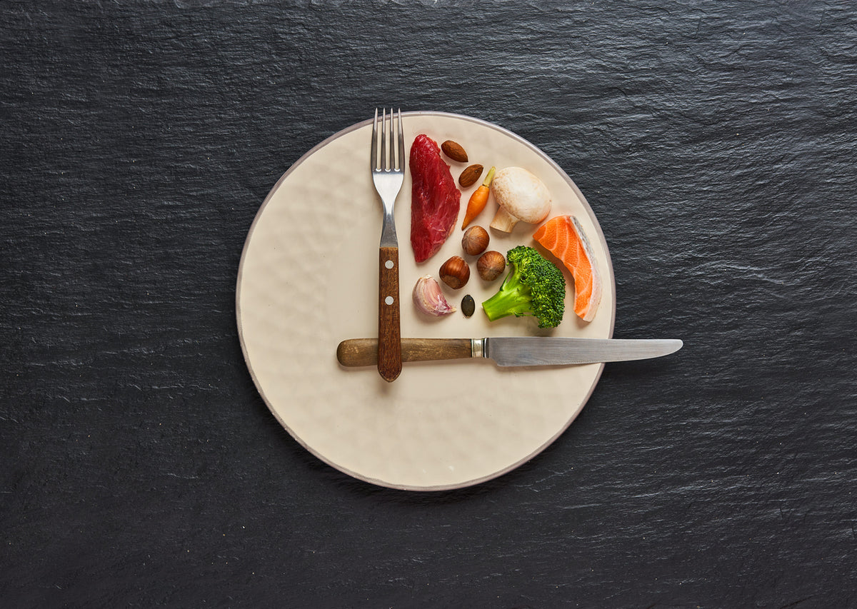 Keto friendly plate with beef, salmon, broccoli, mushrooms and nuts arranged on a dark surface with a knife and fork.