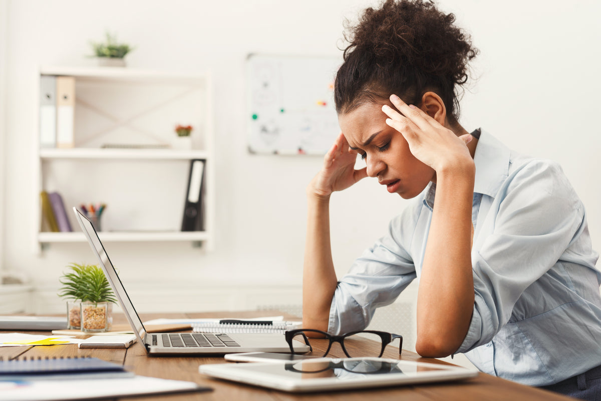 Woman at a bright office desk clutching her temples while working on a laptop.