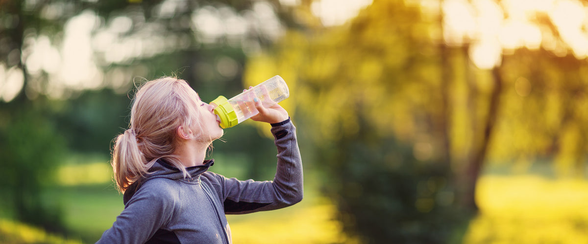 Young woman wearing a gray top drinks from a transparent water bottle with a lime green lid in a sunlit park.